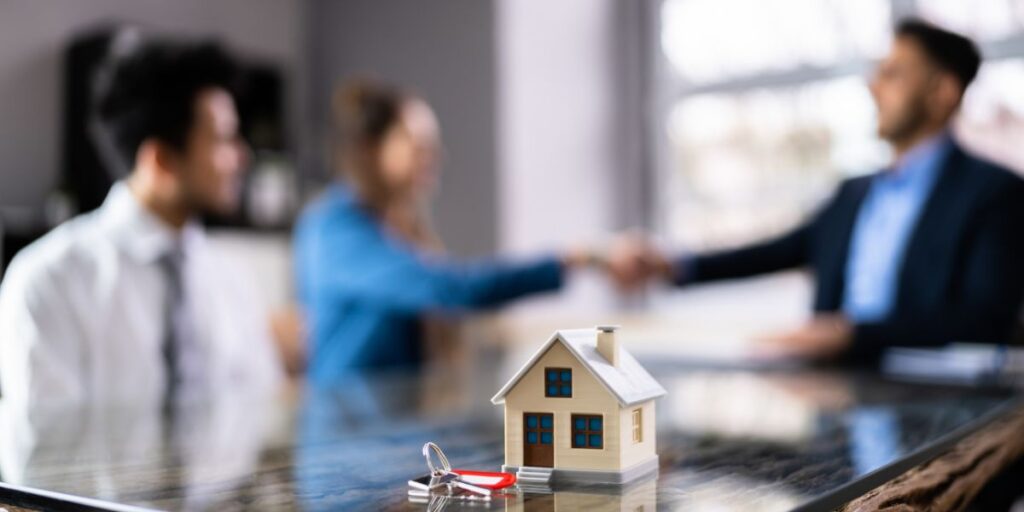 Two blurred figures shaking hands across a table with a miniature house and keys in the foreground, representing a real estate closing or agreement