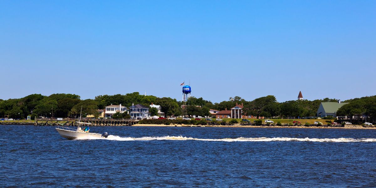 Small motorboat speeding across water near the Southport waterfront with a blue water tower and shoreline buildings in the background.