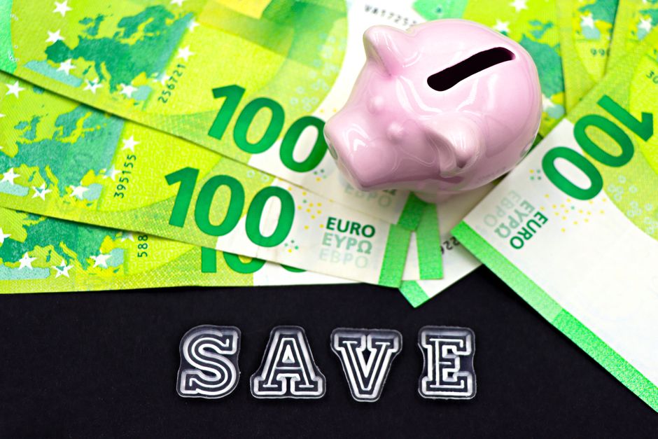 A close-up, minimalist shot of a hand pulling a dollar bill from a white, house-shaped piggy bank, symbolizing the various costs that reduce the final profit from a home sale.