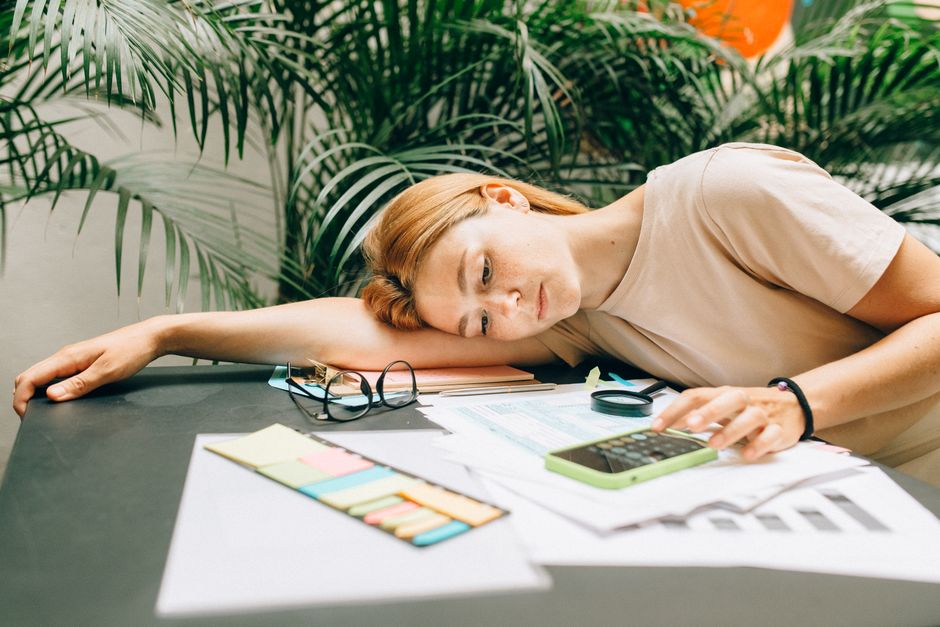 Woman sits at her sunlit kitchen table, looking concerned as she reviews a stack of bills and uses a calculator, represent...